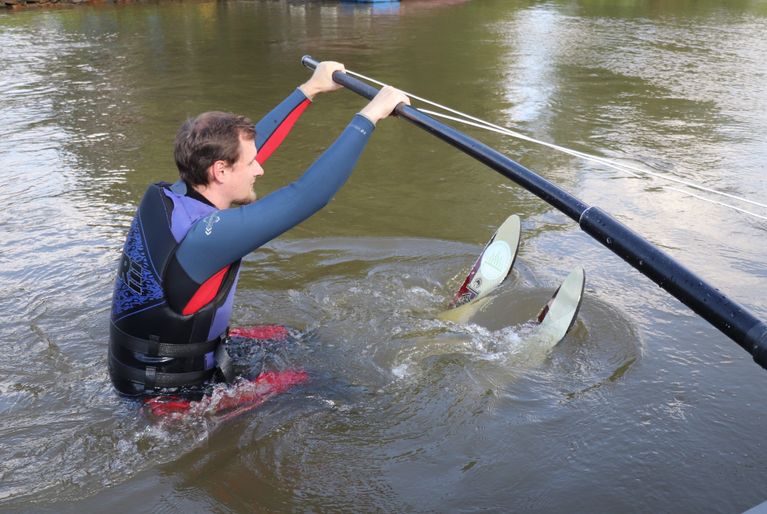 Junger Mann beim Schnupperfahren. Er hat einen Neoprenanzug mit Weste an und hält sich an einer am Boot befästigten Stange fest.